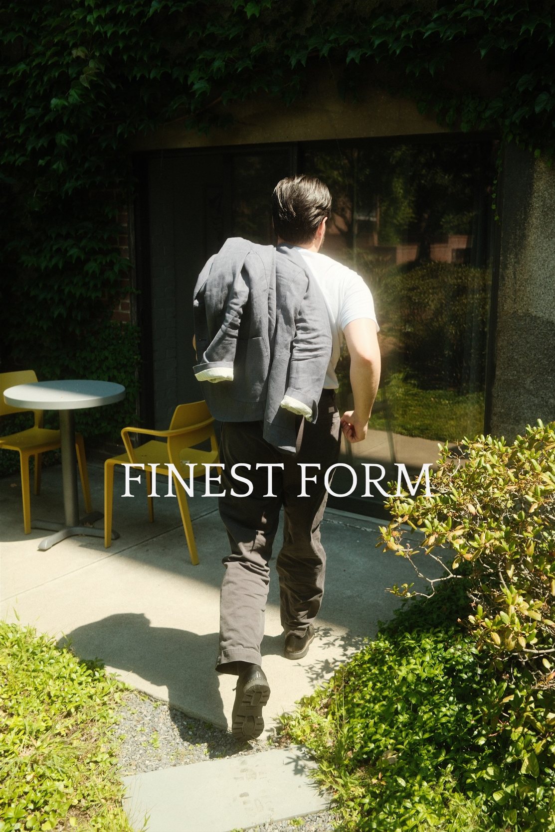 A man walks along a sunlit garden path, a grey linen jacket over his shoulder.